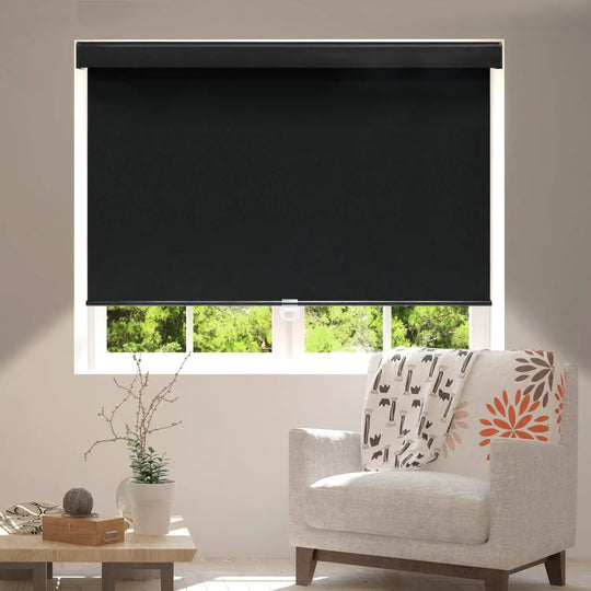 Black blackout roller shade partly lowered over living-room window beside upholstered chair